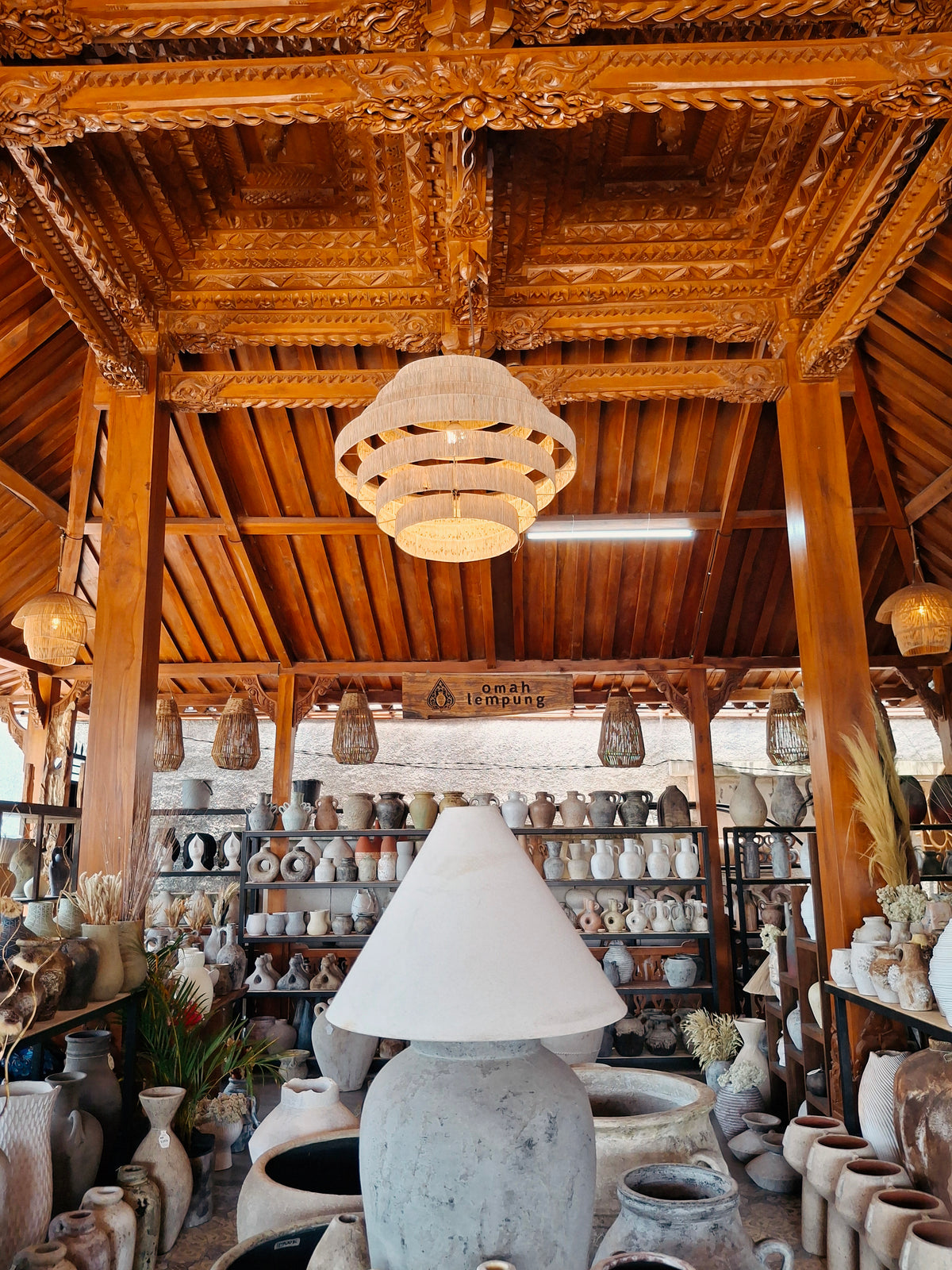 Carved timber roof of the Omah Lempung joglo with a woven pendant lamp and terracotta lamps
